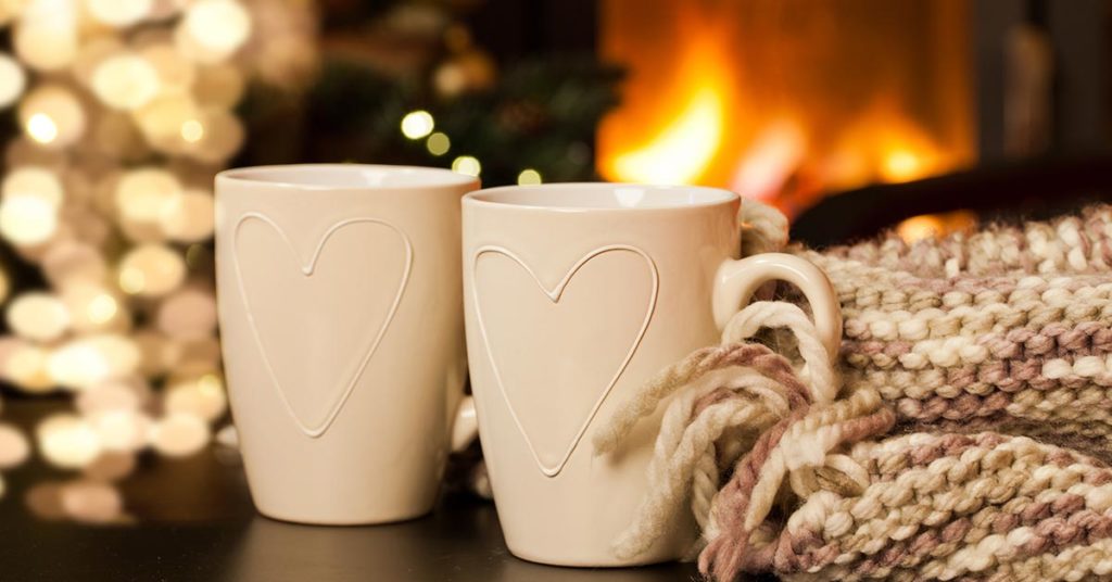 Zwei Tassen mit jeweils einem Herz neben einem Schal und vor einem Weihnachtsbaum und einem Kamin