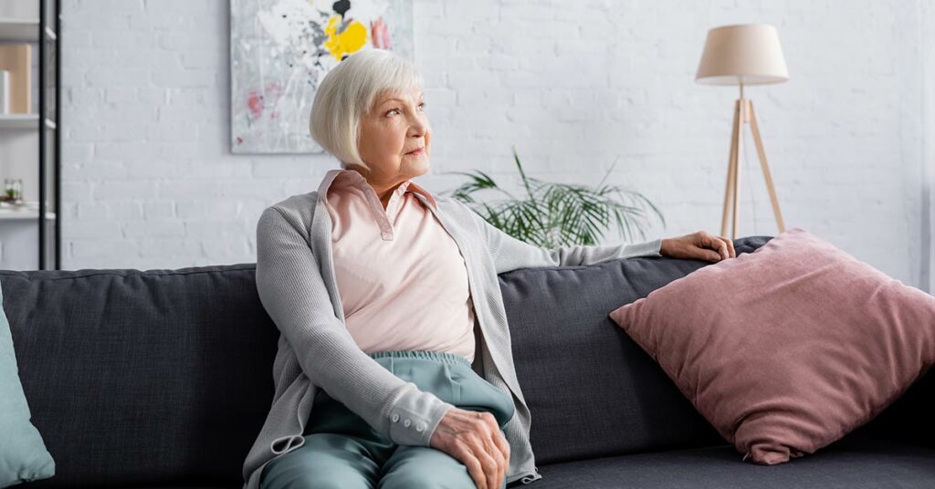 Eine Seniorin sitzt auf ihrem Sofa in ihrem Wohnzimmer, im Hintergrund hängt ein modernes Gemälde, daneben stehen eine Topfpflanze, eine Stehlampe und ein Regal | Wohnen im Alter