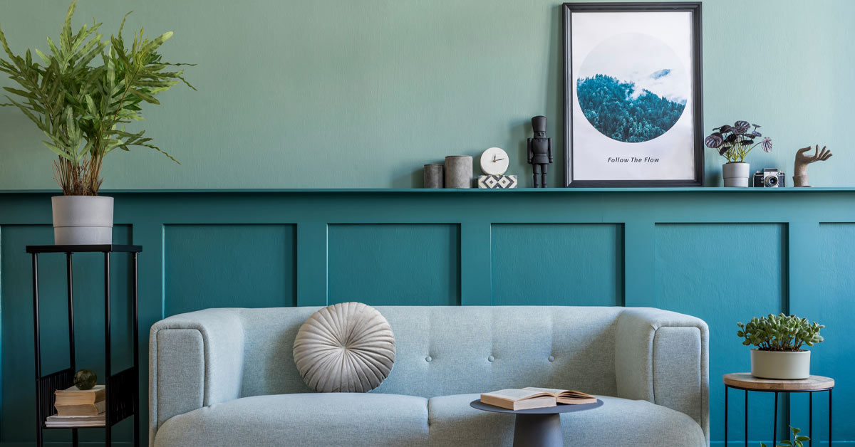 Ein mit Home Staging hübsch eingerichtetes Wohnzimmer mit einem eleganten Sofa, mit einem Beistelltisch mit einem Buch, mit Pflanzen rechts und links des Sofas sowie weiteren schlichten Deko-Elementen | Home Staging