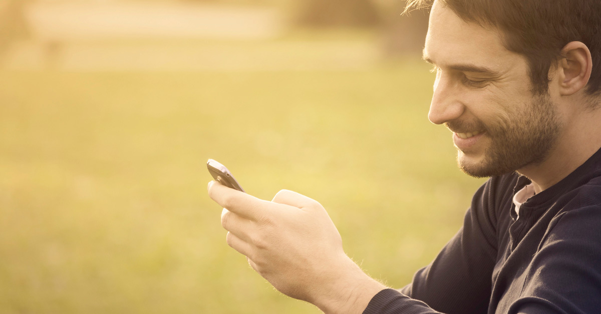 Ein junger Mann sitzt in der Natur und freut sich, am Handy eine Immobilie zum Kauf gefunden zu haben | Immobilienkauf