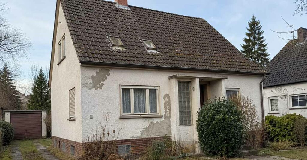 Ein älteres Einfamilienhaus mit verwitterter Fassade und sanierungsbedürftigem Dach steht in einer Wohnstraße und symbolisiert die Entscheidung zwischen Modernisierung und Verkauf. I Sanierungspflicht beim Verkaufen